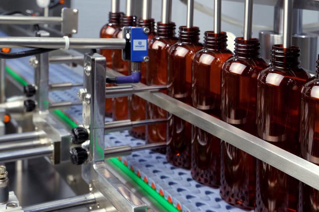 Amber plastic bottles held in a row on a conveyor with fill nozzles lowered into their necks