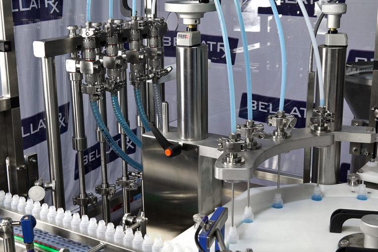 Small dropper bottles on a rotary starwheel being filled with blue liquid through needle nozzles